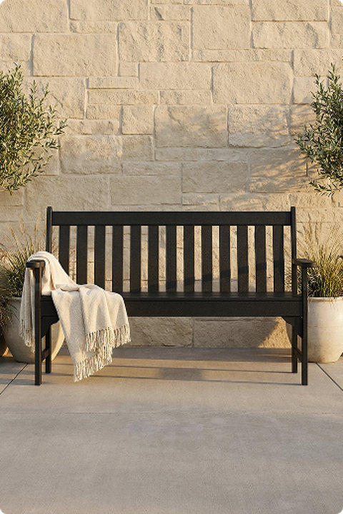 A black POLYWOOD outdoor bench with a draped throw blanket against a stone wall, flanked by potted plants on a patio