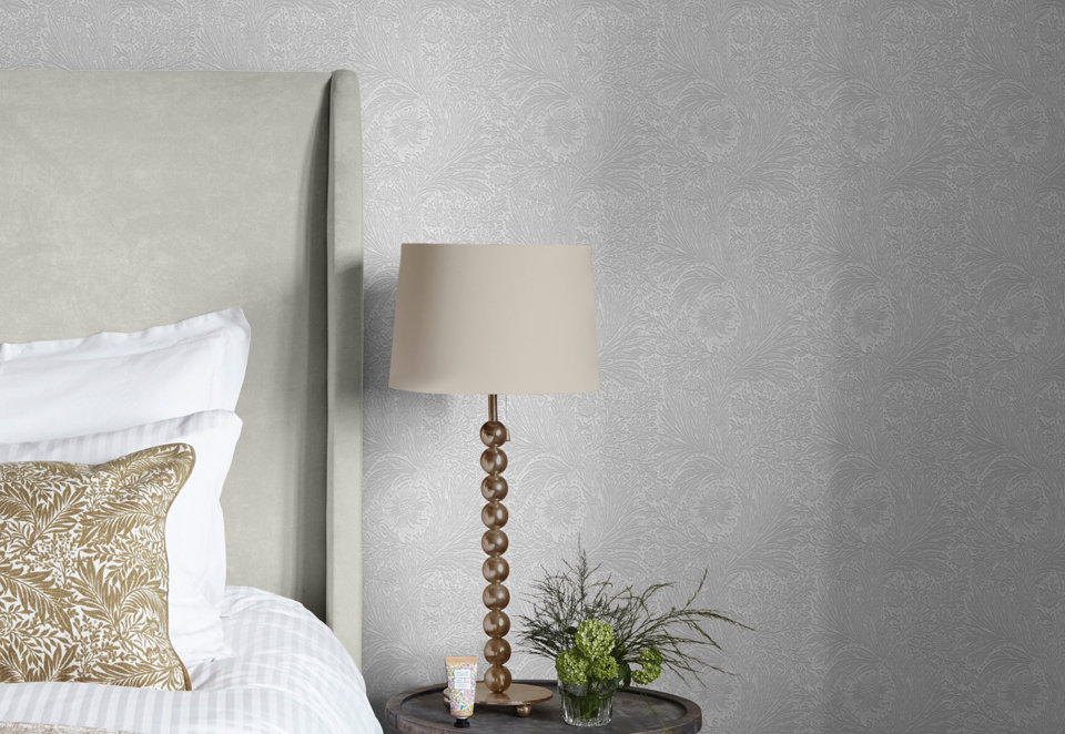 Light gray floral patterned wallpaper behind a bedroom nightstand featuring a beaded table lamp, potted greenery, and a small tube of hand cream.