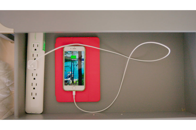 Close-up of an opened nighstand drawer with a smartphone and charging cord plugged into a white power strip.