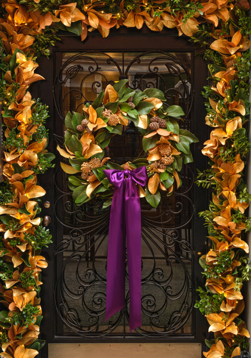 A front door decorated for the holidays with a magnolia and pinecone wreath featuring a long purple ribbon, framed by matching magnolia and greenery garlands with warm string lights.