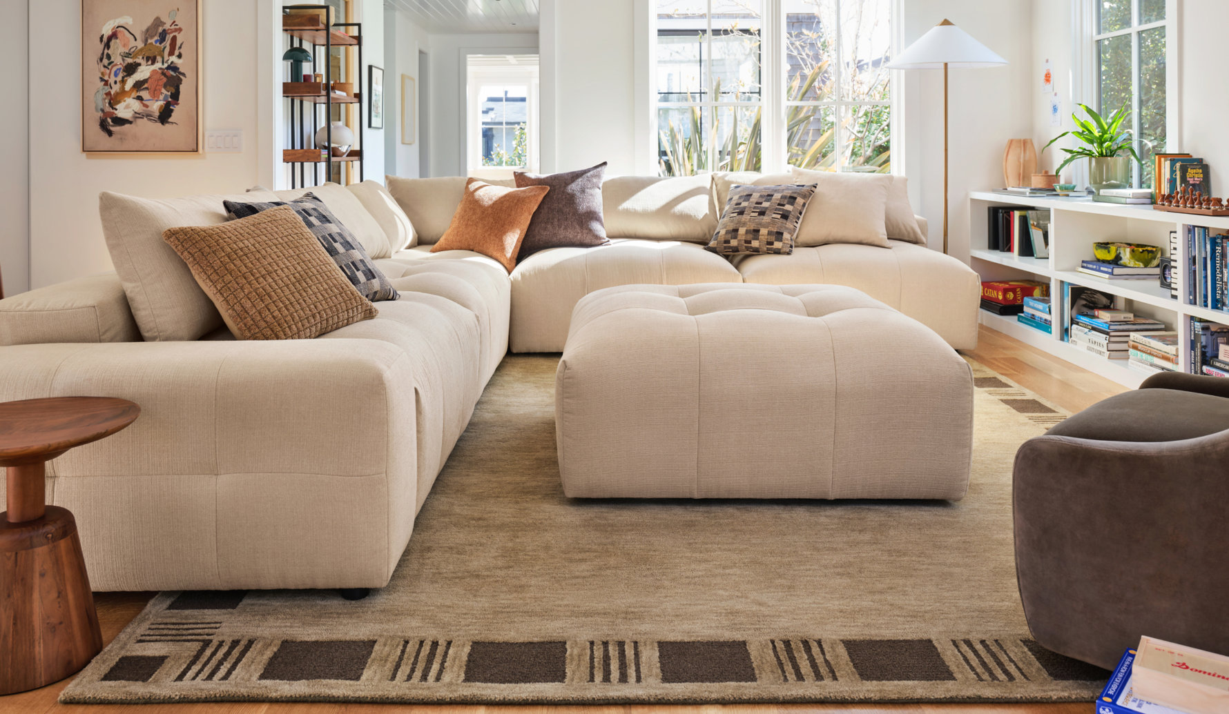 Modern beige sectional sofa with a matching tufted ottoman in a bright, sunlit living room featuring a geometric area rug and a white bookshelf.