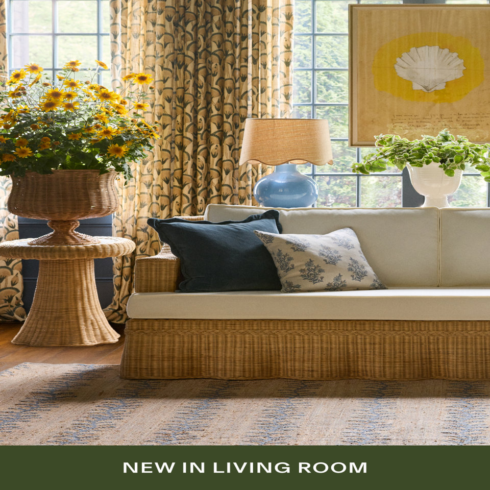 An elegant living room featuring a wicker-base sofa with cream cushions, navy and floral pillows, a blue ceramic lamp with woven shade, yellow sunflowers in a rattan planter, and botanical curtains. Text reads 'NEW IN LIVING ROOM'.