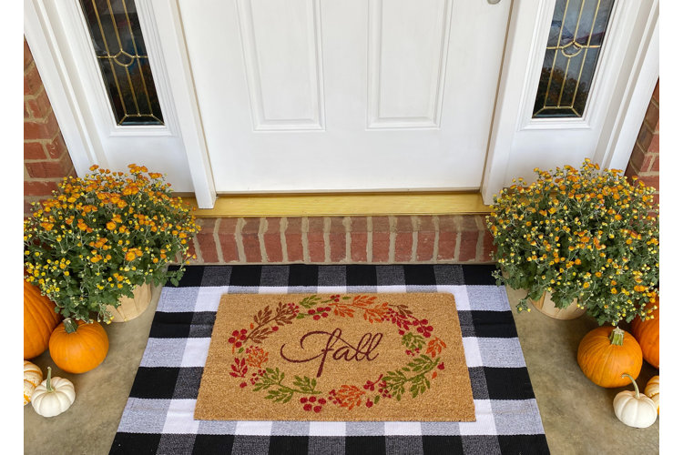 A front porch doorway with a black buffalo check rug and a fall-themed doormat with mums and pumpkins on either side.
