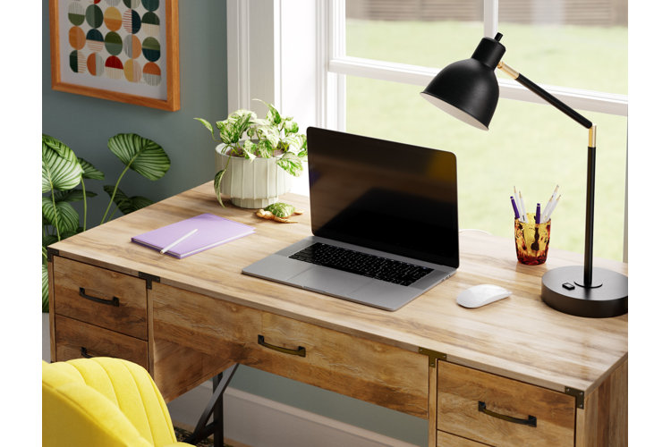 Close up of a wide wooden desk that has room for a potted plant, a notebook, a laptop, office supplies, and a desk lamp.