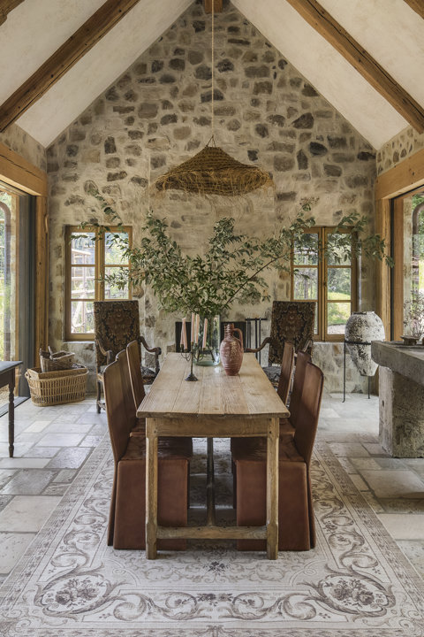 Salle à manger rustique avec une longue table en bois, des chaises en cuir à housse, des murs en pierre et un plafond voûté aux poutres apparentes.