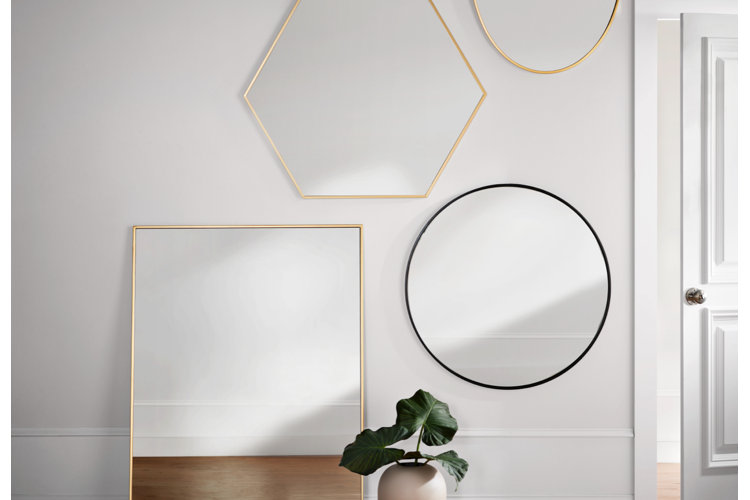 A collection of geometric wall mirrors with minimalist thin metal frames, including a large gold square floor mirror, a gold hexagon mirror, and a thin black round mirror hung on a light gray wall