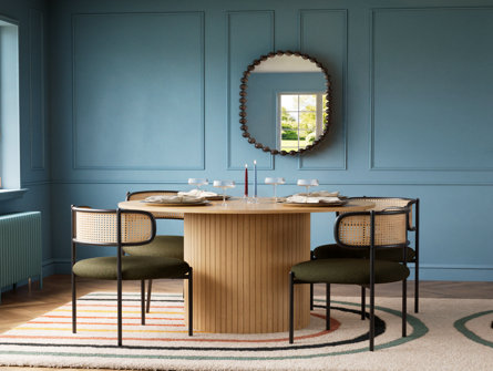 Round wood dining table with ribbed pedestal base and cane-back chairs with dark green seats in a blue panelled room with a decorative mirror.