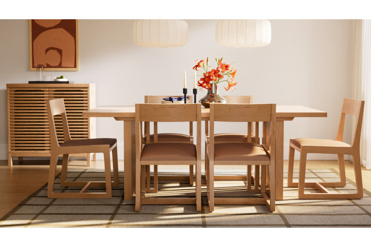 A bright, modern dining room featuring a long light wood dining table with six matching wood chairs that have brown cushioned seats. The table is decorated with a silver vase holding vibrant orange lilies, a pair of black candlesticks, and some folded napkins. In the background, a light wood slatted credenza stands against the wall below an abstract terracotta painting, and two large white paper lantern pendant lamps hang from above. The entire setup rests on a grid-patterned area rug.
