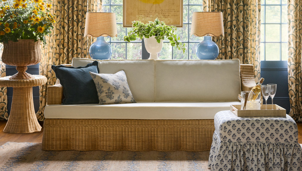 Woven rattan sofa with off-white cushions, matching ottoman, and blue decorative accents in a sunlit room.