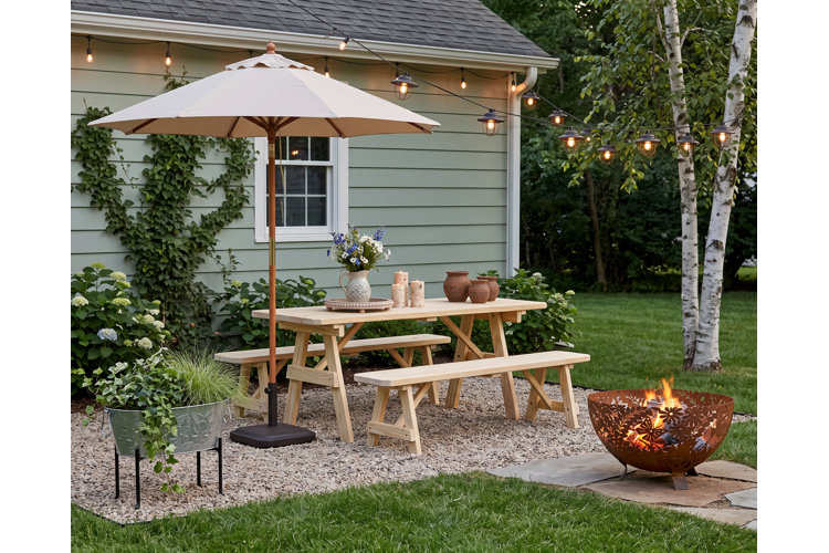 gravel patio area with a wooden picnic table, outdoor umbrella, and metal fire pit on pavers.