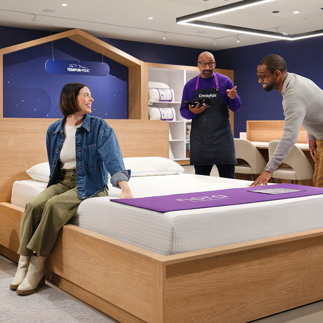 A woman sits on a mattress while a Wayfair associate helps a man try the bed.
