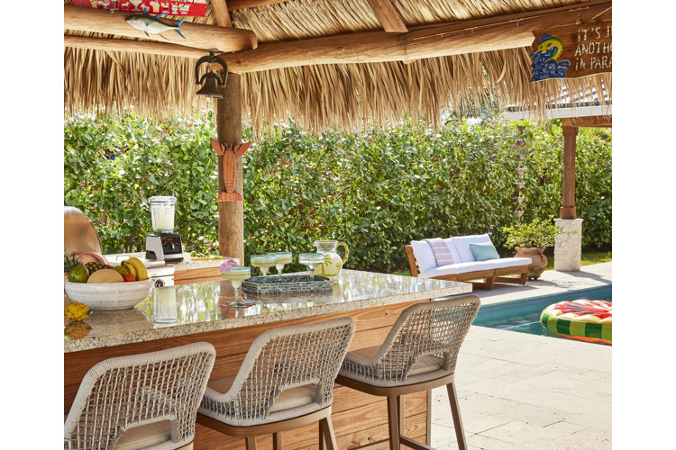 outdoor kitchen island with three rattan bar stools and a tray of margaritas beneath a seagrass cabana and beside a pool.
