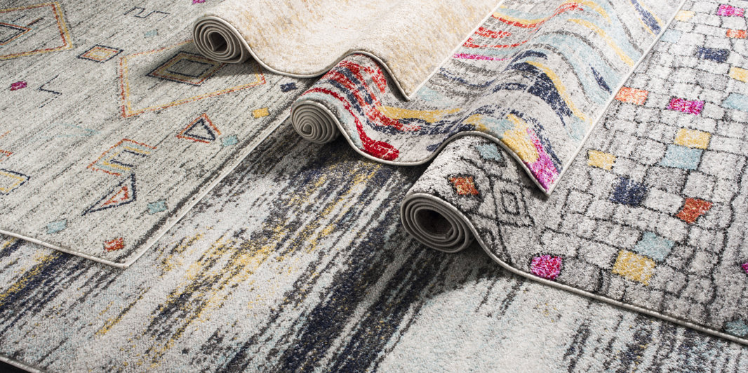 Trois tapis de zone aux motifs abstraits, géométriques et usés dans des tons de gris, bleu, jaune et rouge, partiellement roulés pour mettre en valeur leurs textures et leurs motifs.