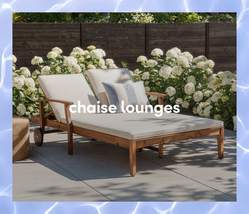 A wooden outdoor double chaise lounge with white cushions and a striped pillow in front of white flowering bushes.