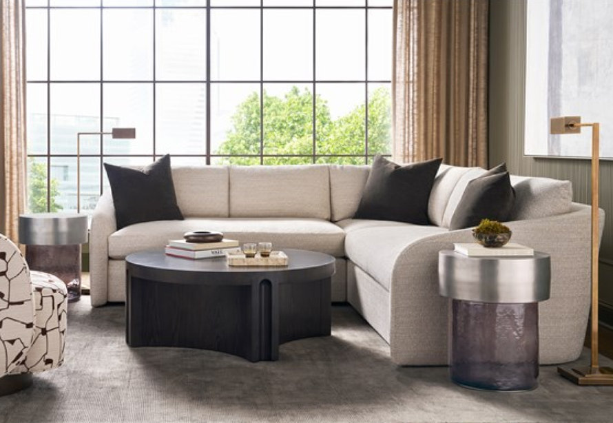 A beige sectional sofa accented with dark gray pillows sits in a sunlit living room featuring a dark wood circular coffee table and modern metallic side tables.