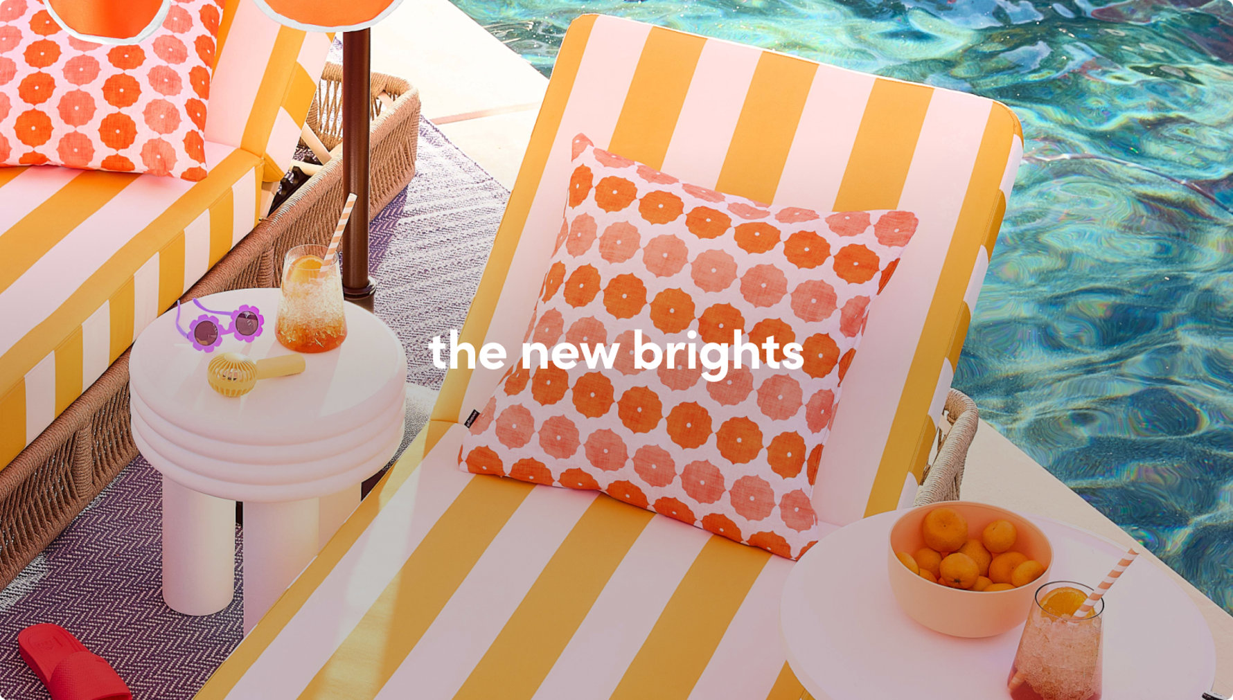 Close-up of a yellow-striped lounge chair with an orange patterned pillow next to a poolside drink and sunglasses.