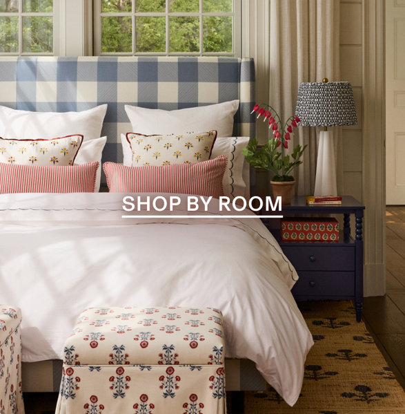 Cozy bedroom featuring a blue and white buffalo check headboard, white bedding with red and floral accent pillows, and a blue nightstand with a patterned lamp, with the text SHOP BY ROOM overlaid.