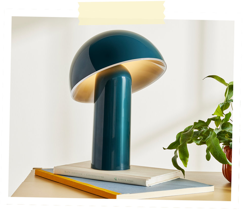 Une lampe de table brillante en forme de champignon de couleur sarcelle éclairant une pile de livres à côté d'une plante verte.