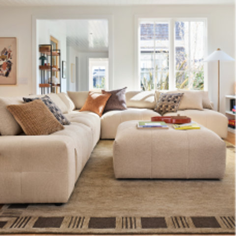 Beige sectional sofa with a matching ottoman and decorative accent pillows in a modern living room setting.
