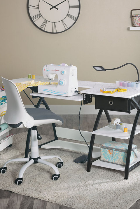 White sewing desk with a black metal frame, a white rolling chair, and a sewing machine set up on a craft room rug under a large wall clock.