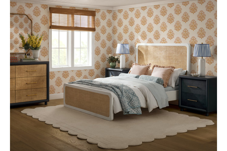 A modern bedroom with orange and white patterned wallpaper a white bed with woven headboard blue white and orange bedding black nightstands and a white area rug with scalloped edges under a queen size bed