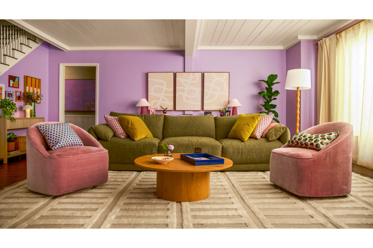 A vibrant, eclectic living room with lavender walls and a white paneled ceiling, featuring a large olive green sofa, two dusty pink velvet swivel chairs, a round oak coffee table, and a triptych of abstract line art
