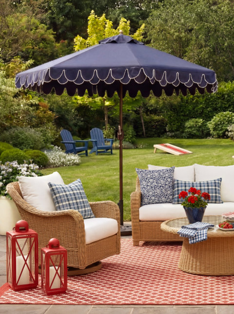 Outdoor patio seating area featuring wicker furniture with white cushions, a navy scalloped umbrella, a red coral outdoor rug, red lanterns, and a lush garden in the background