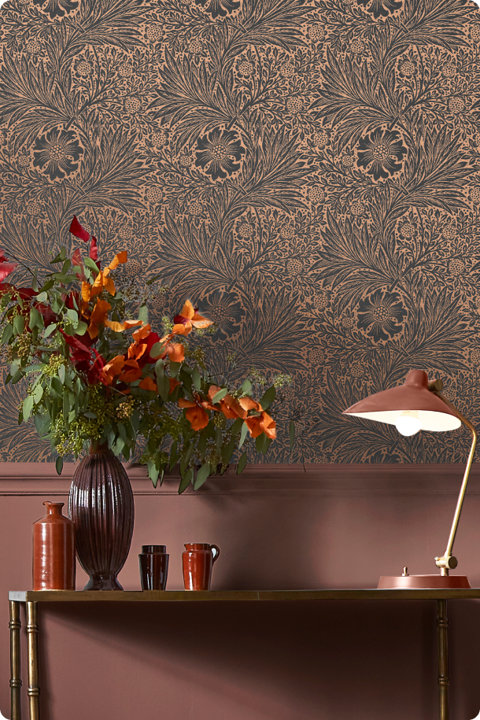 Intricate charcoal floral wallpaper against a warm terracotta wall with a decorative console table featuring a vase of orange foliage and a brass desk lamp.