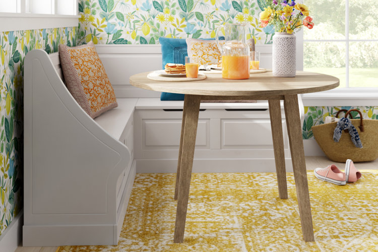 breakfast nook with a white L-shaped bench, round wood table, yellow rug, and floral throw pillows