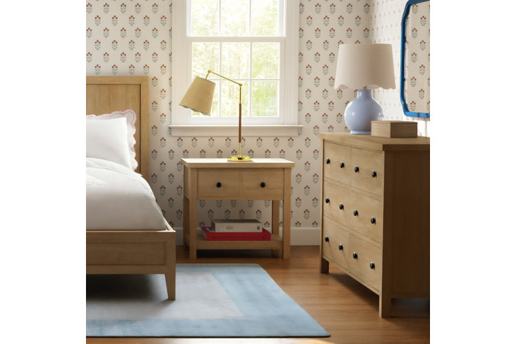 A bright and charming bedroom featuring a light wood panel bed made up with crisp white bedding, scalloped pillows, and a red striped accent pillow. On either side, matching light wood nightstands hold brass reading lamps with scalloped tan shades, set in front of windows with small floral patterned wallpaper. On the right, a matching light wood dresser is styled with a soft blue lamp, and the entire bed set rests on a light blue area rug with a darker blue border.