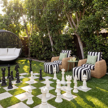Jardin aménagé avec un échiquier géant sur gazon, deux fauteuils en osier à rayures avec poufs assortis, une table d'appoint verte et un fauteuil suspendu en forme d'œuf sous des guirlandes lumineuses.