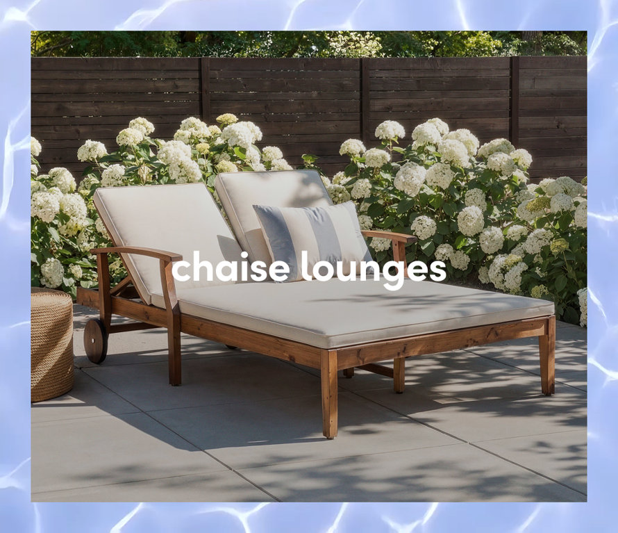 A wooden outdoor double chaise lounge with white cushions and a striped pillow in front of white flowering bushes.