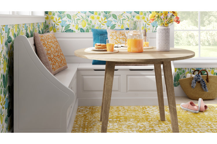 A cheerful, sunlit breakfast nook featuring a round light wood dining table paired with a built-in white corner bench. The bench is cozy with patterned orange, yellow, and blue throw pillows, set against a lively lemon and botanical patterned wallpaper. On the table, a breakfast spread includes pancakes, glasses of orange juice, a pitcher, and a textured white vase with bright yellow wildflowers. A vibrant yellow pattern area rug covers the floor, with a pair of pink sneakers and a woven tote bag sitting nearby.
