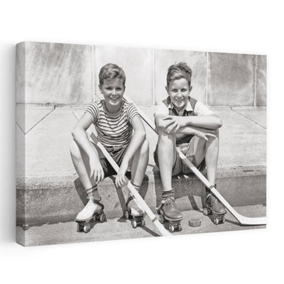 Années 1930, deux garçons souriants, joueurs de hockey de rue assis sur un trottoir, portant des patins à roulettes en métal tenant des bâtons de hockey, rondelle couchée, rue, rétro