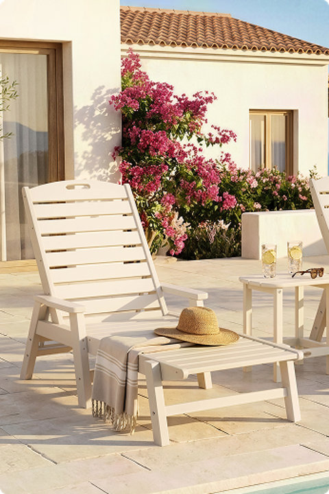 Sand-colored POLYWOOD chaise lounge chair on a sunny patio beside a white house with blooming pink flowers and a side table holding drinks