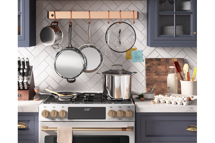 Rustic kitchen with a copper hanging pot rack. Stainelss steel range with a large pot and pan. White counter with a knife block and wood cutting board all below blue cabinets