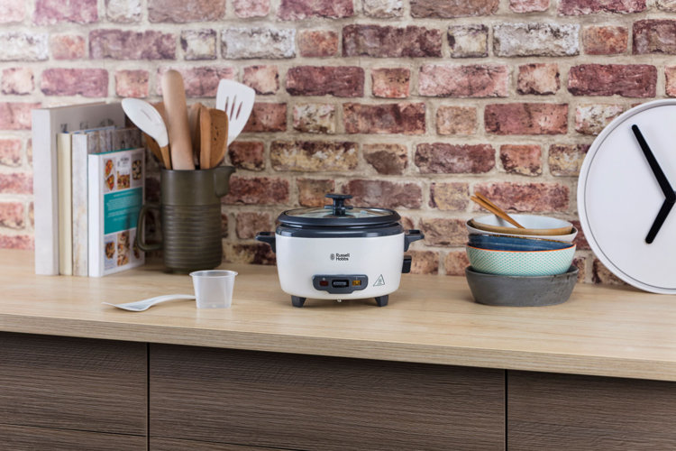 a rice cooker on a wood counter. there is a stack of bowls next to it, and a cup of utensils.