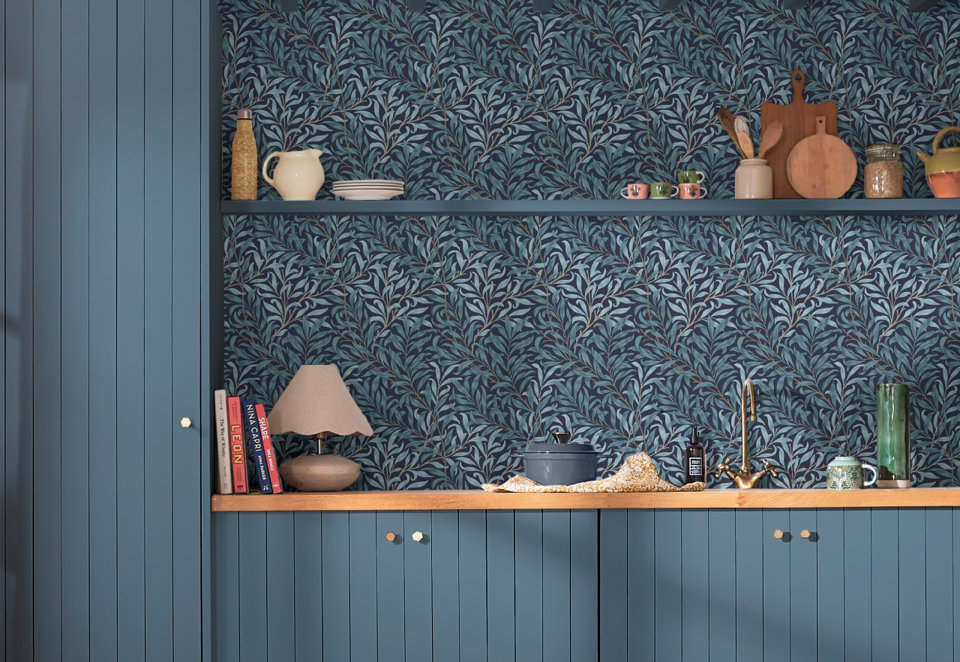 Blue kitchen cabinetry featuring a natural wood countertop, a dark wall with teal botanical leaf wallpaper, and open shelving styled with ceramics, wooden boards, and kitchen accessories.
