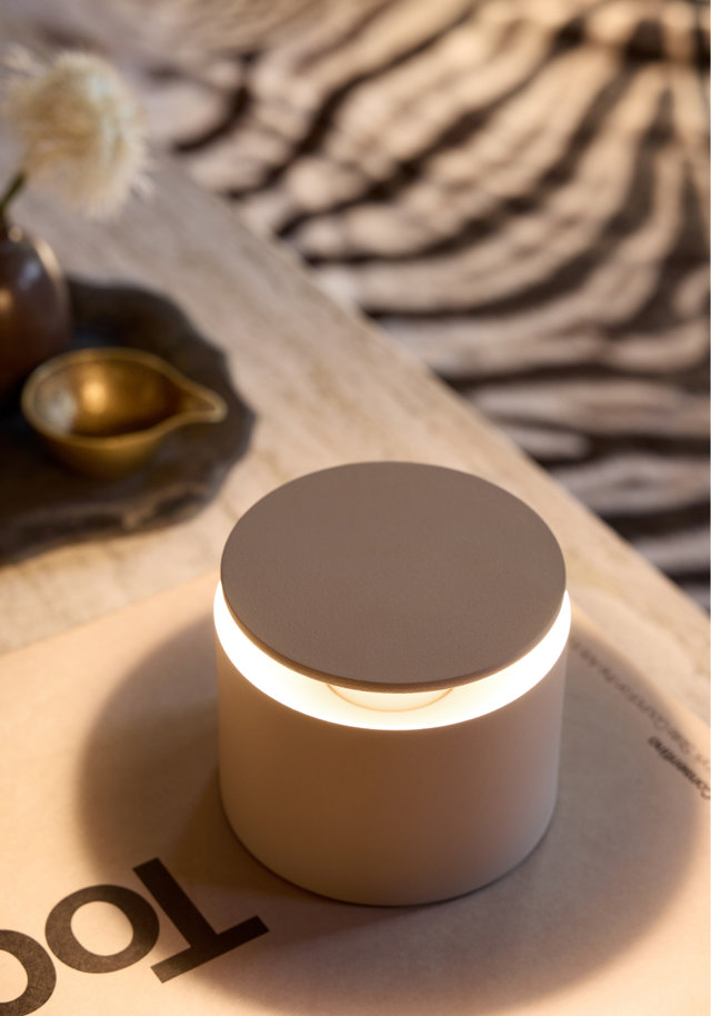 A modern, cylindrical white table lamp with a glowing ring of light sits on a coffee table next to a decorative bowl and a zebra-print rug in the background.