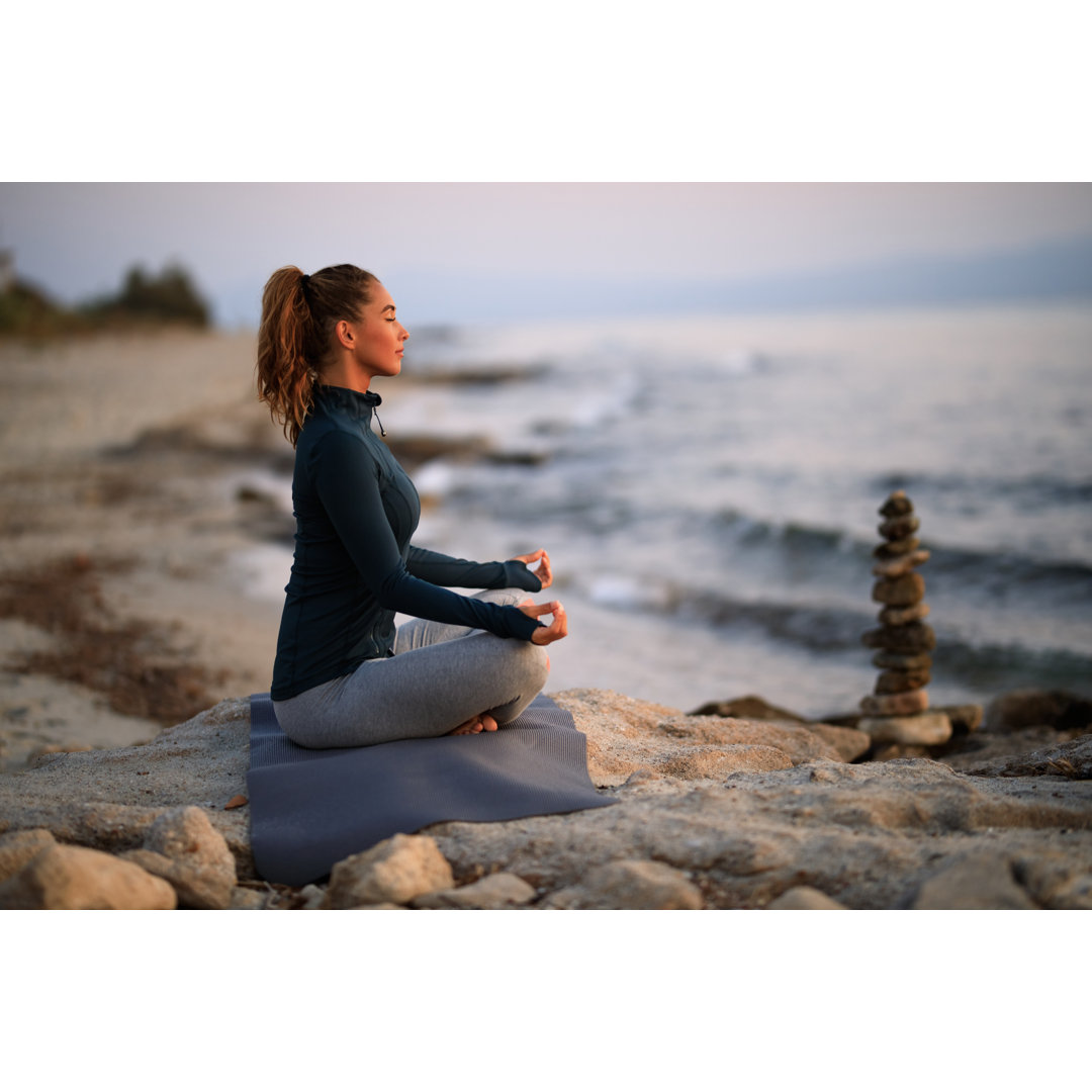 Wandbild Junge Frau meditiert in Lotus Position auf einem Felsen am Strand