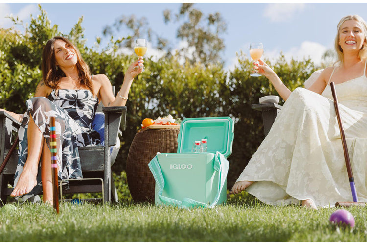 two women sitting in lawn chairs outside on the grass with a mint green small hard-sided igloo travel cooler in between them with a beverage in the cooler and each with a drink in their hand