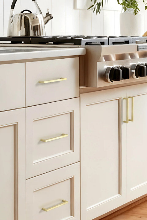 Modern matte gold cabinet pulls installed on cream-colored kitchen cabinetry next to a stainless steel stove.
