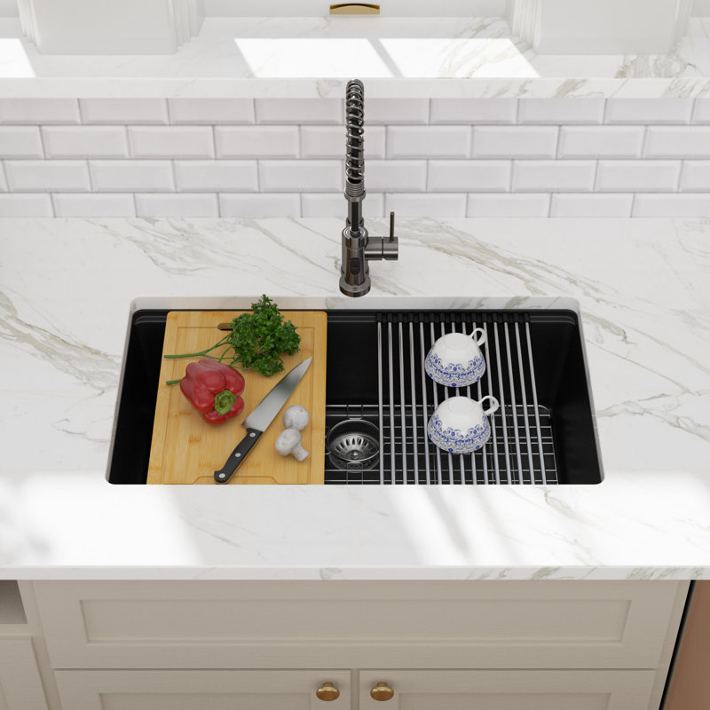 Black quartz undermount workstation kitchen sink with a wooden cutting board and roll-up drying rack in a marble countertop.