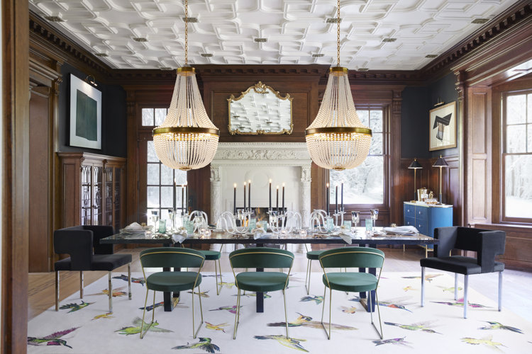 A dining room with wood paneled walls, two crystal chandeliers, a rectangular dining table, and green velvet dining chairs.