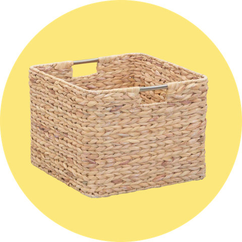 A woven seagrass storage basket with white pom-pom trim and carrying handles, on a circular yellow background.