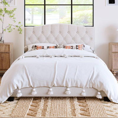 Grand lit plateforme tapissier beige, avec détails en losanges et pieds de selle courbés courbés et ornement