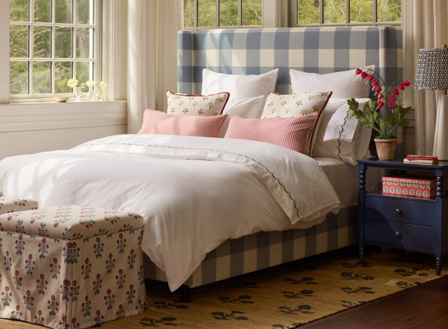 Bedroom featuring a blue and white buffalo check upholstered headboard and a white duvet with scalloped edges, styled with floral and red striped accent pillows, a floral patterned bench, and a dark blue nightstand.
