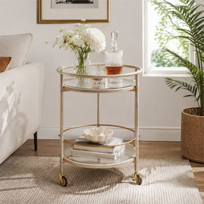 A round mobile bar cart with tempered glass shelves and gold accents; the cabinets have an aged white finish.