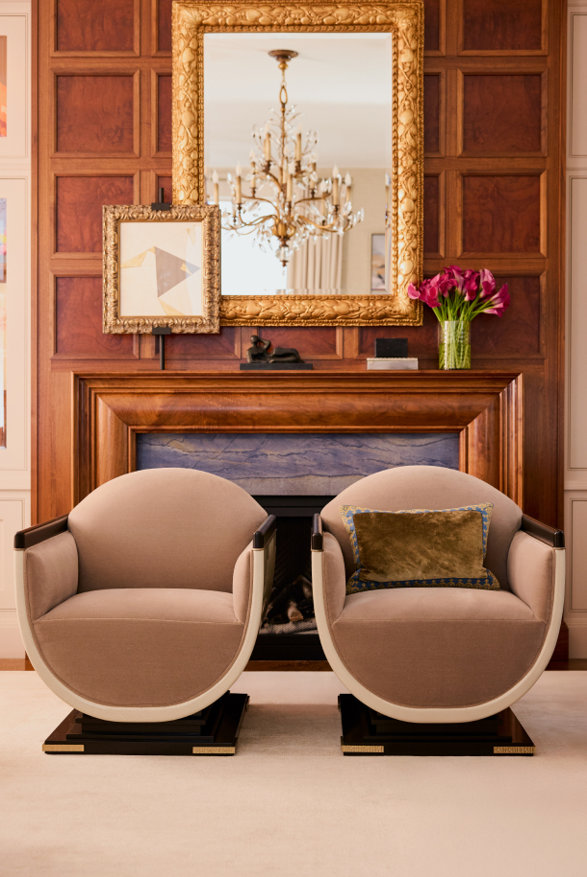 Two rounded armchairs in front of fireplace with a mirror and chandelier above.