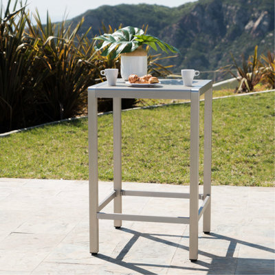 Table de bar extérieure avec structure en aluminium et plateau en verre gris hypoallergénique. Remarque : Ceci contrevient aux instructions, mais est inclus à titre d'exemple de ce qu'il ne faut pas faire. En réalité, le mot-clé « allergie » est autorisé. Résistante aux intempéries et
